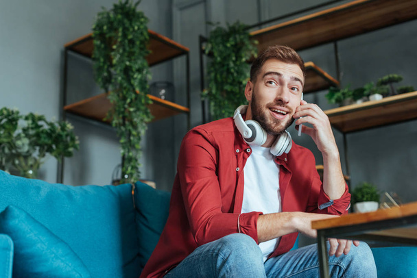 low angle view of happy bearded man in headphones talking on smartphone in living room  - Fotó, kép