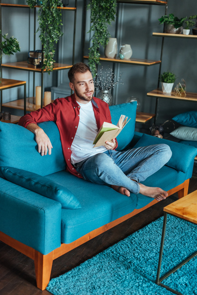 handsome man sitting on sofa and holding book in living room  - Fotó, kép