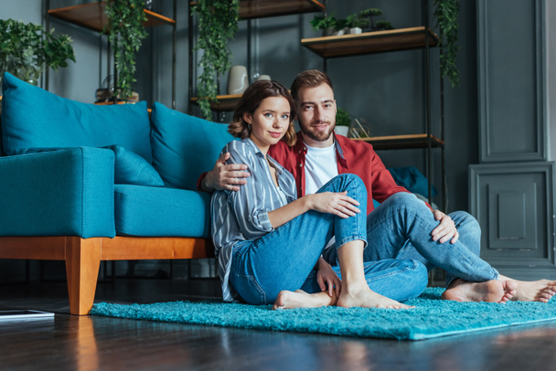 low angle view of cheerful man hugging attractive woman while sitting on carpet at home  - Foto, afbeelding