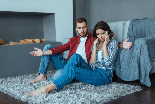 upset man looking at attractive woman talking on smartphone while sitting on carpet  - Foto, Imagem