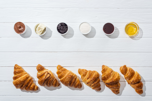 Schüsseln mit Frischkäse, Schokocreme, Sauerrahm, Marmelade und Honig in der Nähe von frischen Croissants auf Weiß  - Foto, Bild