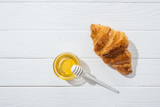 Draufsicht auf leckeres und frisches Croissant in der Nähe von Glasschale mit Honig und Honiglöffel auf weißer Oberfläche  - Foto, Bild