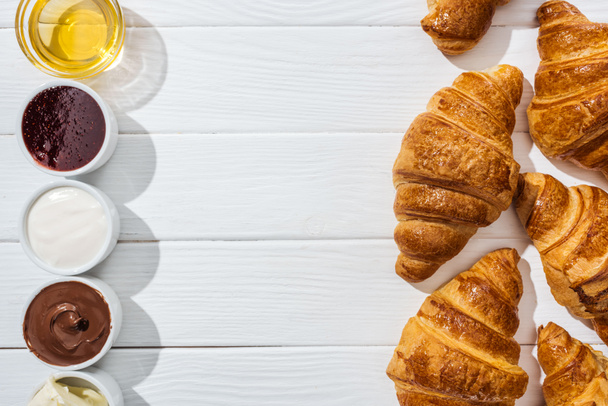 top view of tasty croissants near bowls with cream cheese, chocolate cream, sour cream, jam and honey on white - Foto, afbeelding