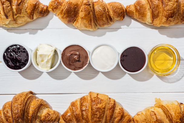 Blick von oben auf köstliche Croissants in der Nähe von Schüsseln mit Frischkäse, Schokoladencreme, saurer Sahne, Marmelade und Honig auf weißem Hintergrund - Foto, Bild