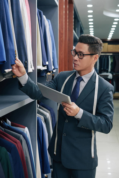 Вьетнамский помощник в магазине мужской одежды с цифровыми планшетами, проверяющими кнопки на куртках на стойках
 - Фото, изображение