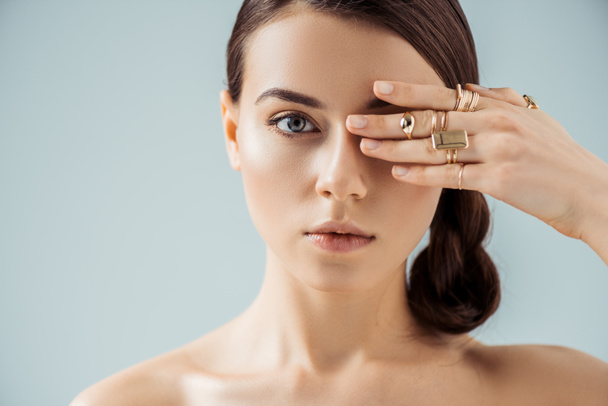 young nude woman with shiny makeup and golden rings hiding eye behind hand isolated on grey - Foto, Imagem