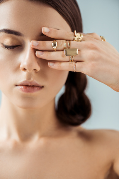 young naked woman with shiny makeup and closed eyes in golden rings hiding eye behind hand isolated on grey - Foto, Imagem