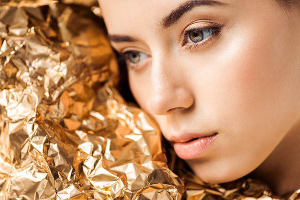 Portret van jonge vrouw met glanzende make-up in de buurt van gouden folie op zoek weg - Foto, afbeelding