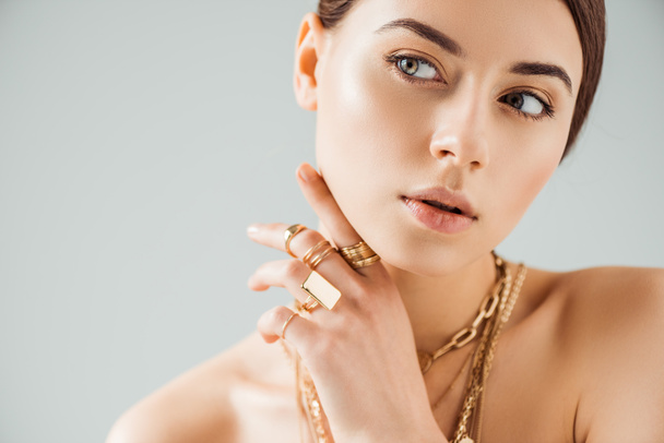 νεαρή γυμνή γυναίκα με χρυσά δαχτυλίδια και περιδέραια κοιτάζει μακριά απομονωμένη στο γκρι - Φωτογραφία, εικόνα