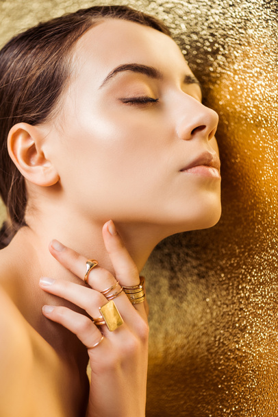 nude young woman with closed eyes, shiny makeup and golden rings on golden textured background - Φωτογραφία, εικόνα