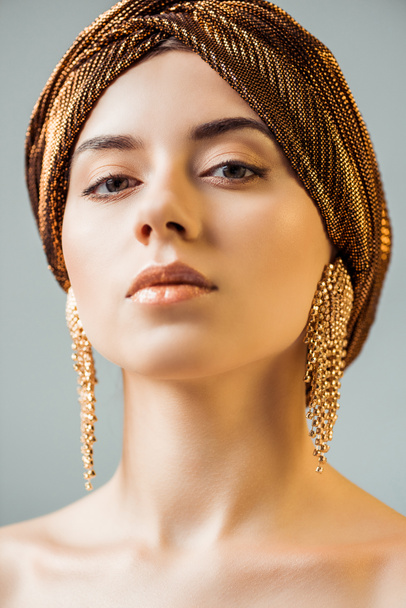 young naked woman with shiny makeup, golden rings in turban looking at camera isolated on grey - Photo, Image