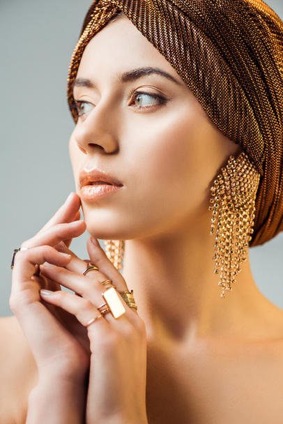 young naked woman with shiny makeup, golden rings and earrings in turban looking away isolated on grey - Foto, imagen
