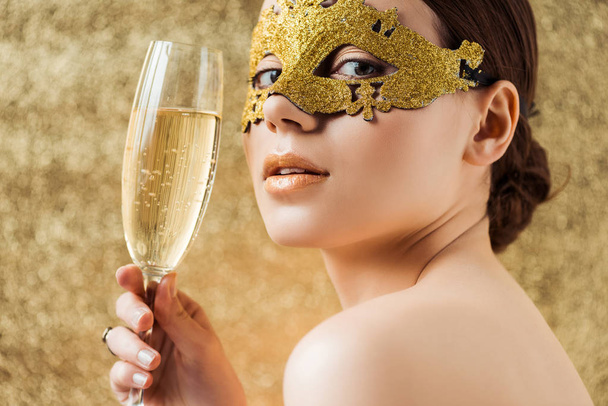 junge nackte Frau in goldener Maske mit glänzendem Make-up und einem Glas Champagner vor der Kamera auf strukturiertem goldenem Hintergrund - Foto, Bild