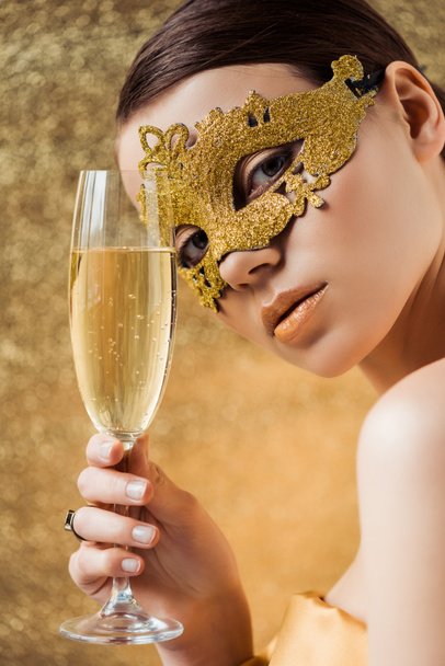 молода жінка в золотій масці з блискучим макіяжем і келихом шампанського дивиться на камеру на золотому фоні
 - Фото, зображення