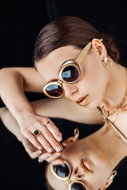 young nude woman in sunglasses, golden jewelry lying on mirror isolated on black - Fotografie, Obrázek