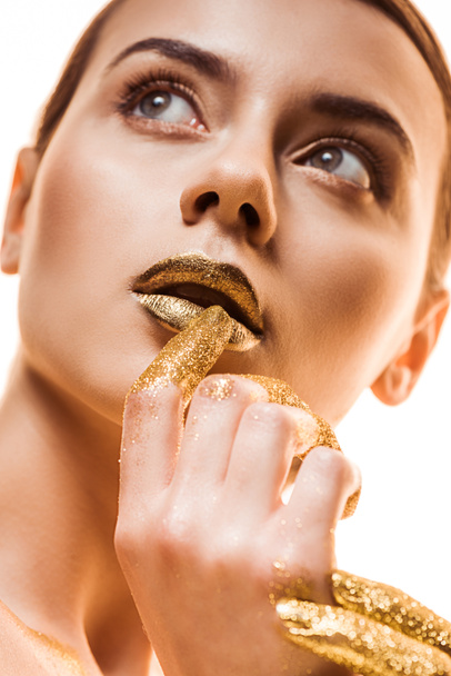 young beautiful woman with golden makeup and sparkles on fingers touching lips and looking away isolated on white - Фото, изображение