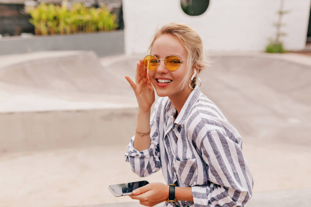 Hippie happy girl with blond hair wearing orange glasses listening music in the headphones and holding smartphone  - Foto, immagini