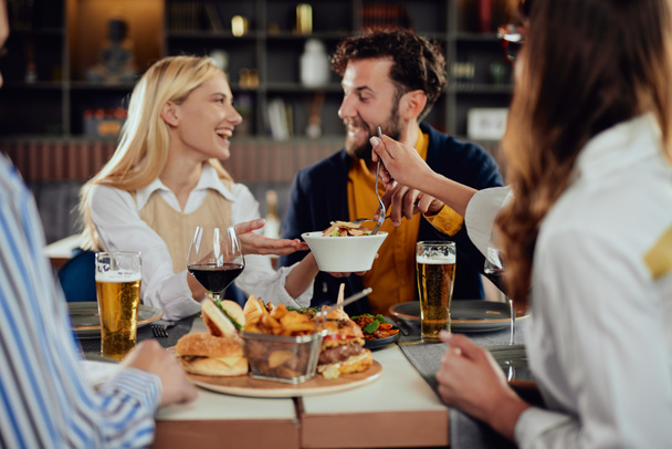 Amigos multiétnicos sentados en el restaurante, bebiendo alcohol, charlando y comiendo hamburguesas para cenar
. - Foto, imagen