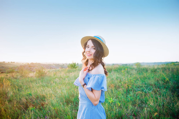 Portrait of young woman in blue romantic dress and straw hat on green meadow enjoying summer sunset. Calm and harmony. Summer vacation,fun, positive mood. Back sun light, sun beams. Copy space. - Fotografie, Obrázek