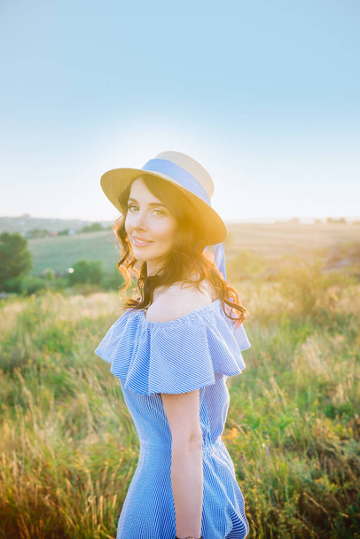 Portrait of young woman in blue romantic dress and straw hat on green meadow enjoying summer sunset. Calm and harmony. Summer vacation,fun, positive mood. Back sun light, sun beams. Copy space. - Zdjęcie, obraz
