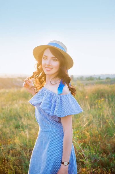 Porträt einer jungen Frau in blauem romantischen Kleid und Strohhut auf einer grünen Wiese, die den sommerlichen Sonnenuntergang genießt. Ruhe und Harmonie. Sommerurlaub, Spaß, positive Stimmung. zurück Sonnenlicht, Sonnenstrahlen. Kopierraum. - Foto, Bild