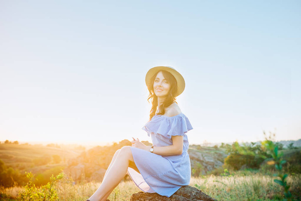 Portrait of young woman in blue romantic dress and straw hat in sun light enjoying summer sunset on nature. Calm and harmony. Summer vacation,fun, positive mood. Back sun light, sun beams. Copy space. - Photo, Image