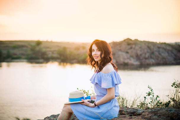 Portrait of young woman in blue romantic dress with hat sitting near river and enjoying sunset evening. Calm and harmony. Summer vacation,fun, positive mood. Back sun light, sun beams. Copy space. - Photo, Image