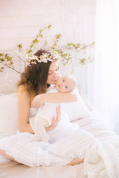 Young cute mom in a wreath of cherry blossoms in a white dress holding a baby in her arms in a spring bedroom with blossoming cherry branches - Fotografie, Obrázek