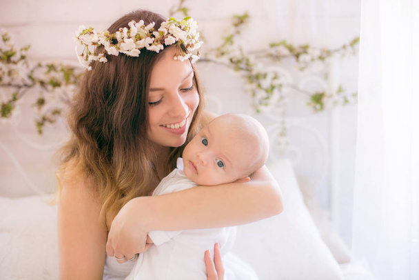 Young cute mom in a wreath of cherry blossoms in a white dress holding a baby in her arms in a spring bedroom with blossoming cherry branches - Foto, Imagen