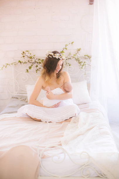 Young cute mom in a wreath of cherry blossoms in a white dress holding a baby in her arms in a spring bedroom with blossoming cherry branches - Fotoğraf, Görsel