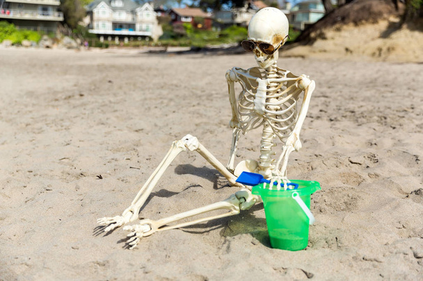Esqueleto senta-se na praia brincando na areia com um balde e pá
 - Foto, Imagem