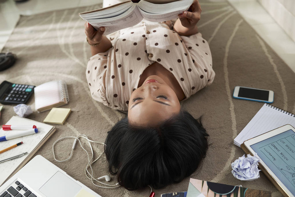 Studentin, die auf dem Boden liegt und sich darauf konzentriert, zu Hause ein Buch zu lesen oder sich auf wichtige Prüfungen vorbereitet - Foto, Bild