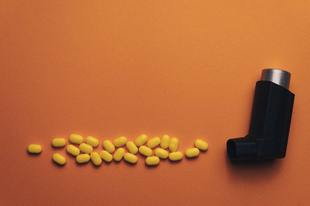 オレンジ色の背景に黄色の錠剤製剤(ビタミン、染料、風味増強剤、栄養補助食品、キャンディスイーツ)によって作られた粒子エアロゾルスペイを持つ黒い喘息吸入器.  - 写真・画像