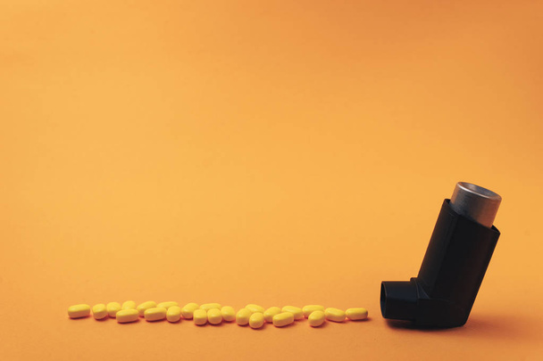 Musta astma-inhalaattori, jossa on keltaisilla pillereillä valmistettuja hiukkasaerosoleja (vitamiineja, väriaineita, arominvahventeita, ravintolisiä, makeisia) oranssilla taustalla
.  - Valokuva, kuva