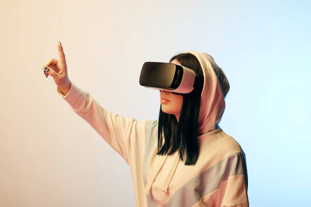 beautiful brunette woman in virtual reality headset pointing with finger on beige and blue  - Photo, Image