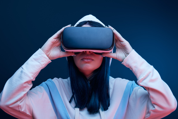 brunette girl in hood touching virtual reality headset on blue - Фото, изображение