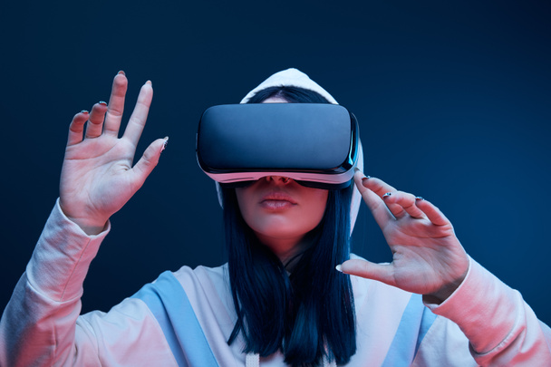 brunetka dziewczyna w kaptur wskazując podczas wirtualny rzeczywistość słuchawki na niebiesko - Zdjęcie, obraz