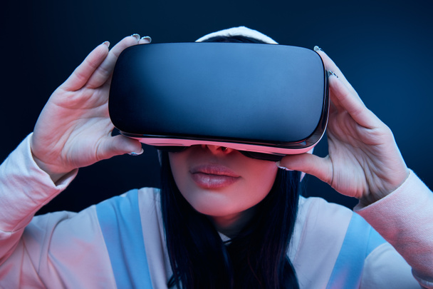 selective focus of brunette woman in hood touching virtual reality headset on blue - Фото, зображення