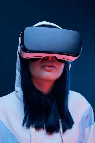 low angle view of brunette young woman in hood using virtual reality headset on blue - Foto, Imagem