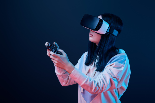 KYIV, UKRAINE - APRIL 5, 2019: Brunette woman holding joystick while playing video game in virtual reality headset on blue  - Фото, изображение