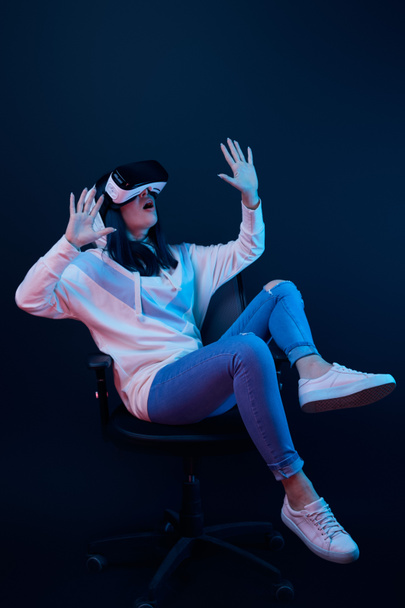 schockierte junge Frau gestikuliert und verwendet Virtual-Reality-Headset, während sie auf Stuhl auf blau sitzt  - Foto, Bild