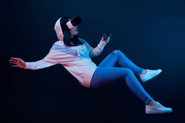 KYIV, UKRAINE - APRIL 5, 2019: Surprised woman in virtual reality headset levitating and holding joystick on blue   - Photo, image