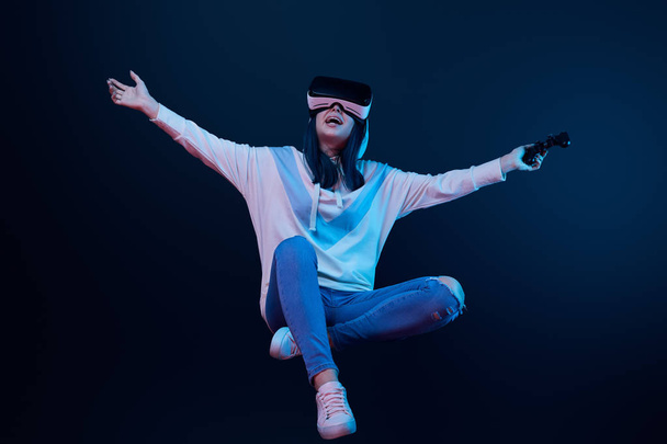 KYIV, UCRANIA - 5 DE ABRIL DE 2019: Mujer feliz con auriculares de realidad virtual levitando y sosteniendo el joystick en azul
   - Foto, Imagen