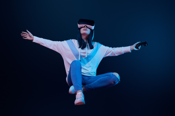 KYIV, UCRANIA - 5 DE ABRIL DE 2019: Mujer joven con auriculares de realidad virtual levitando y sosteniendo el joystick en azul
   - Foto, Imagen