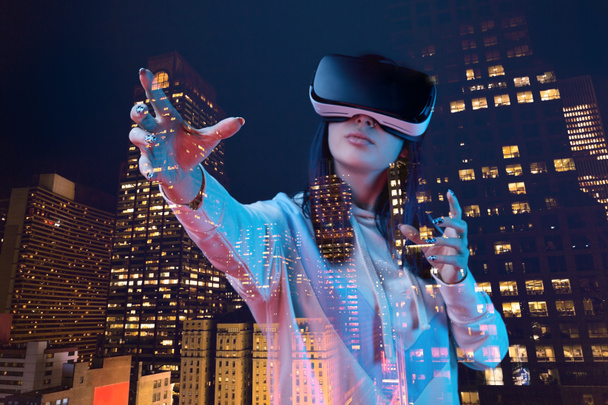 Doppelbelichtung brünettes Mädchen gestikuliert, während es Virtual-Reality-Headset und moderne Stadt mit Wolkenkratzern in der Nacht trägt  - Foto, Bild
