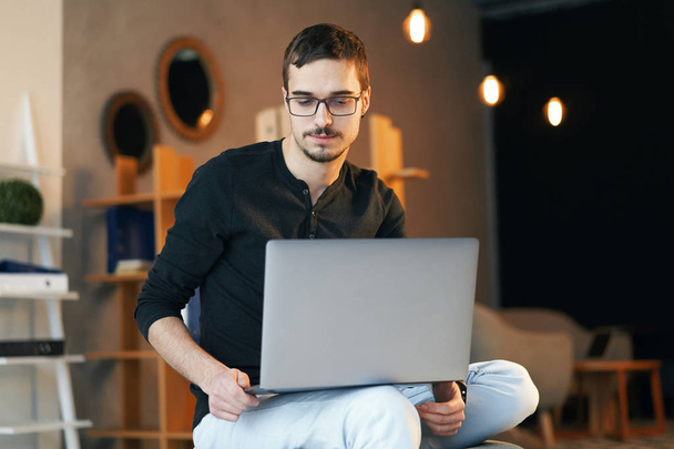 Młody człowiek siedzi z komputerem. Freelancer w okularach z laptopem, kierownik projektu. - Zdjęcie, obraz