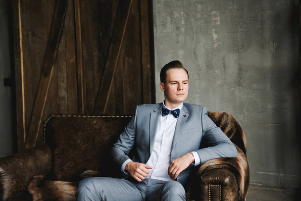 Retrato de um homem. O noivo em um terno cinza, camisa branca e gravata borboleta está sentado em um sofá de couro marrom. Quarto em estilo loft
. - Foto, Imagem