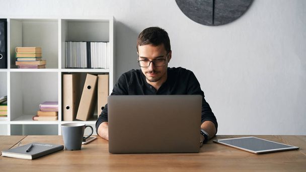 Молодий чоловік працює з комп'ютером, телефоном і планшетом за столом під час пиття кави
 - Фото, зображення
