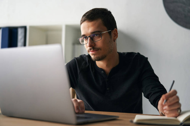 Νεαρός που εργάζεται με υπολογιστή, τηλέφωνο και tablet στο τραπέζι πίνοντας καφέ - Φωτογραφία, εικόνα