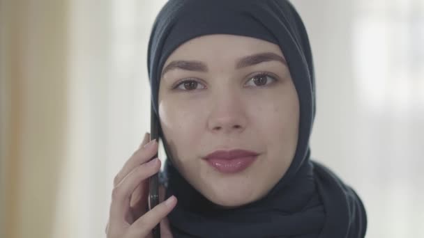 Portrait of a young beautiful eastern woman in the modern Muslim clothes and beautiful black headdress talking by cell phone close up. Young asian Muslim woman in head scarf looking in camera - Séquence, vidéo